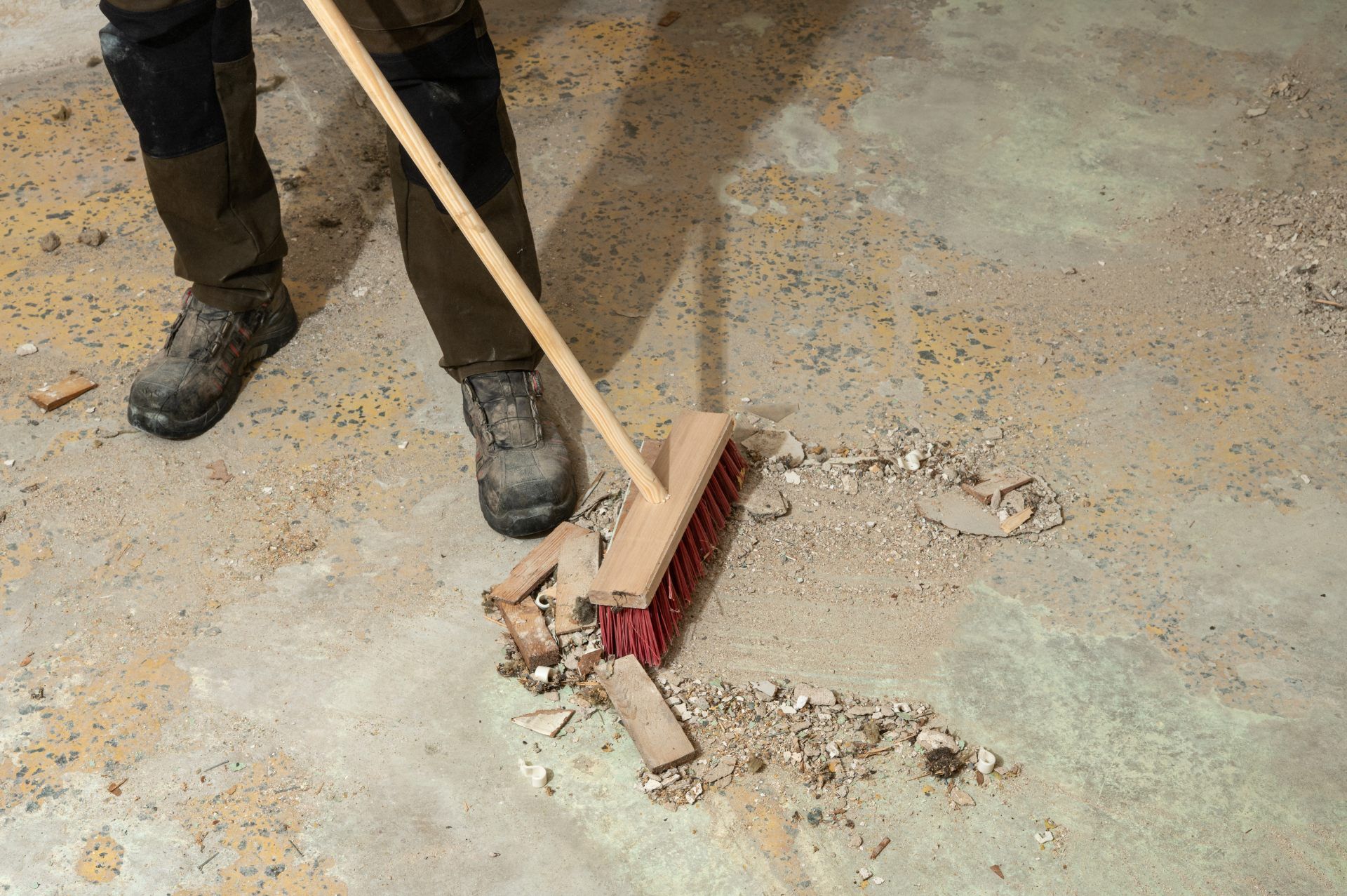 A person wearing work boots uses a push broom to sweep debris off a concrete floor.