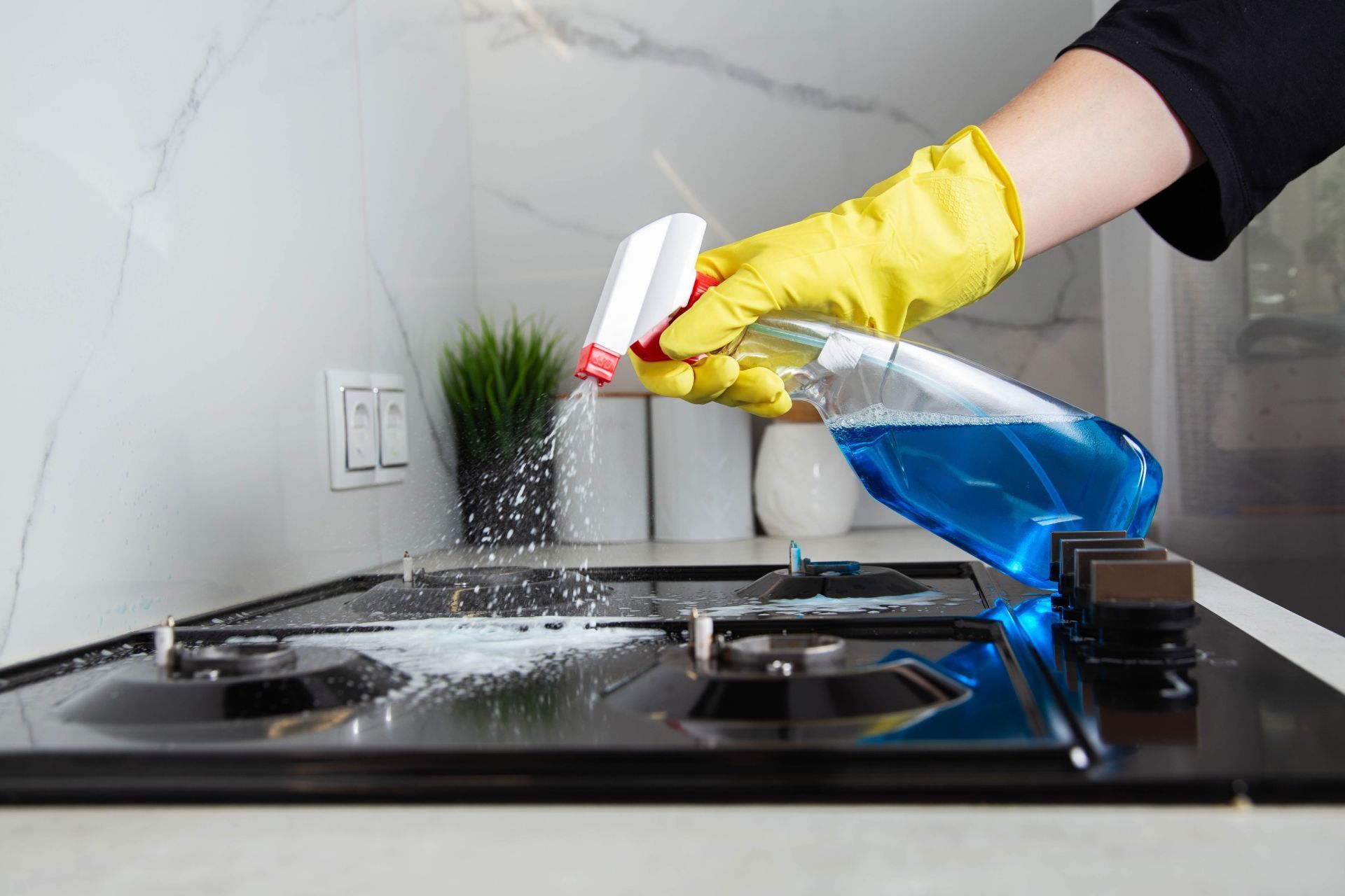 A hand in a yellow rubber glove sprays blue cleaning liquid onto a black kitchen stovetop.