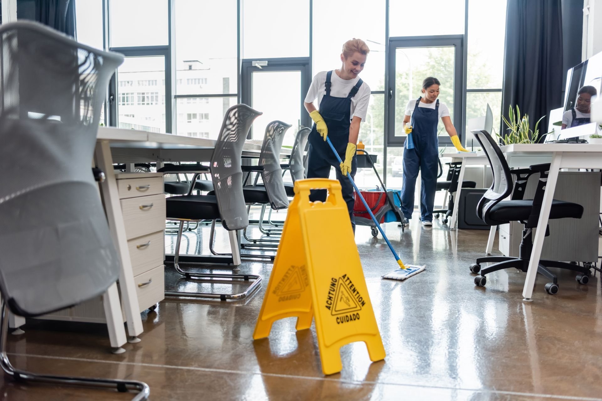 Two professionals in blue overalls clean an office floor behind a yellow caution sign.