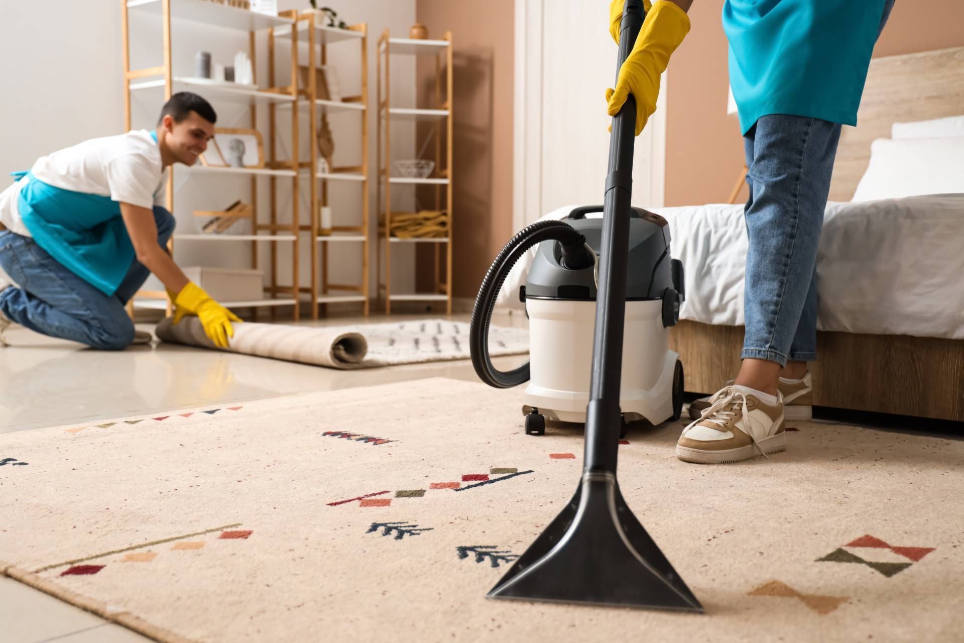 Two professional cleaners in a bright bedroom, one using a carpet cleaner on a rug while another rolls up a second rug.