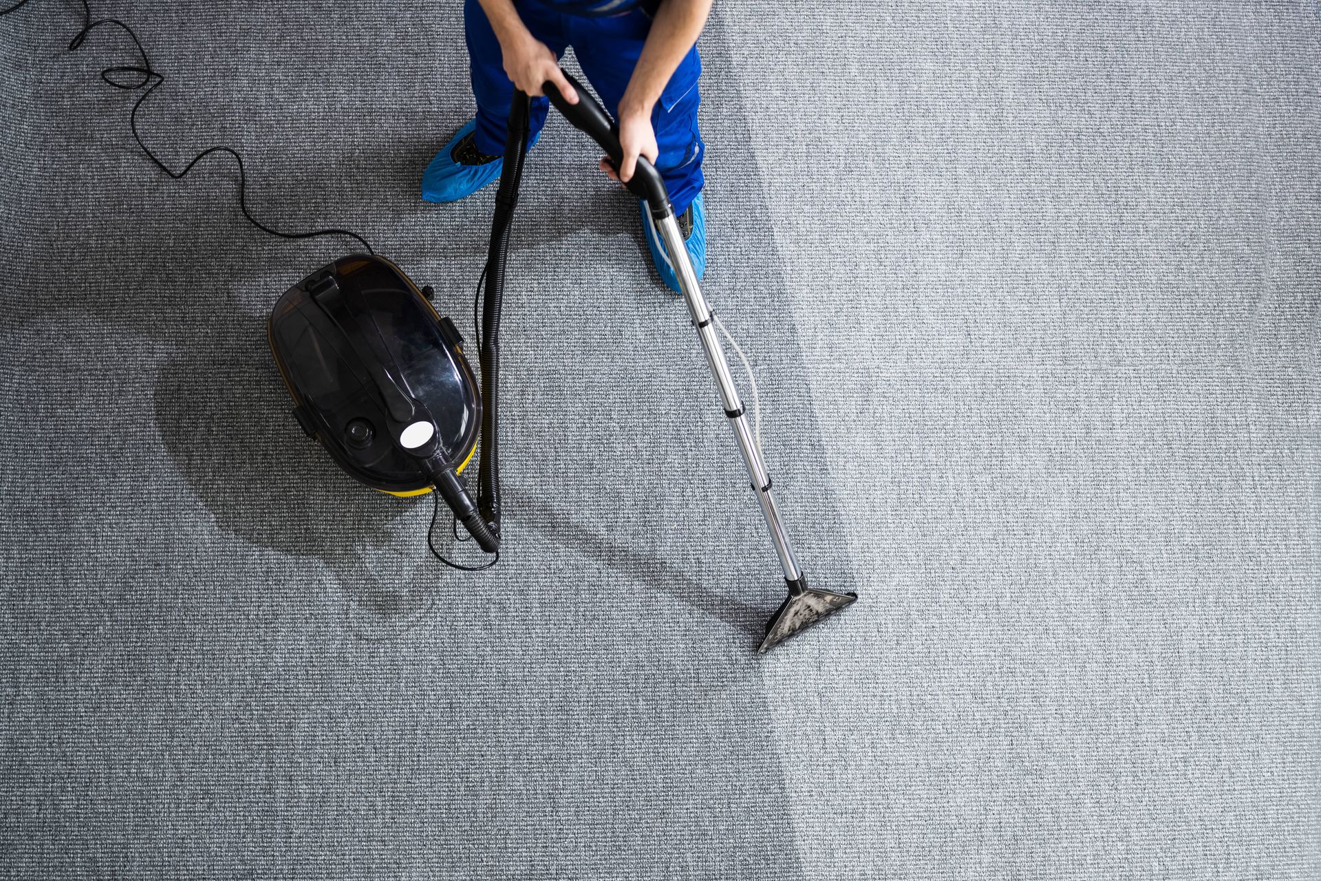 A person uses a black canister vacuum to clean a textured gray carpet, showing a clear path of freshly cleaned surface.