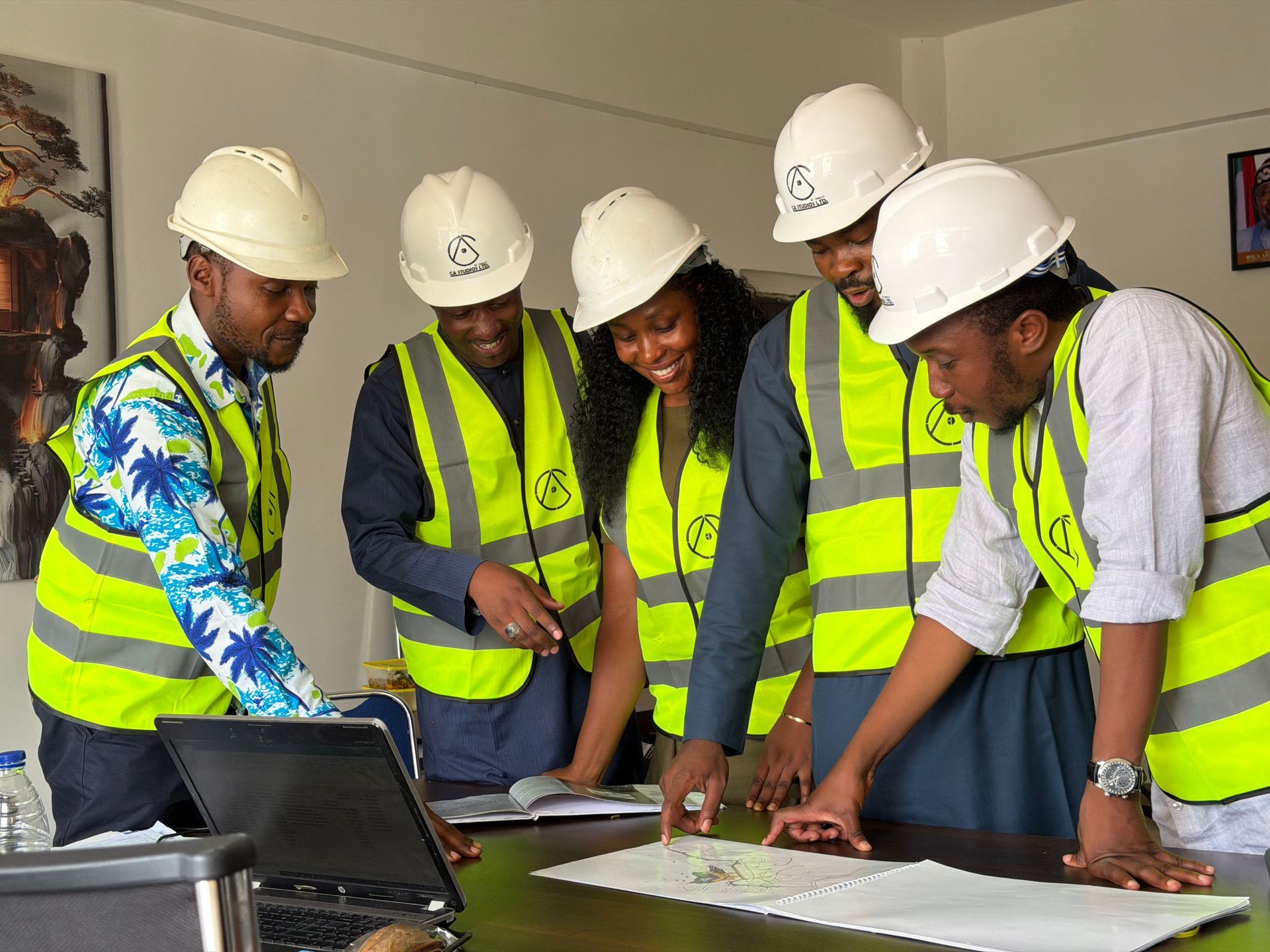 Five construction workers in hard hats review plans around a laptop on a table indoors.