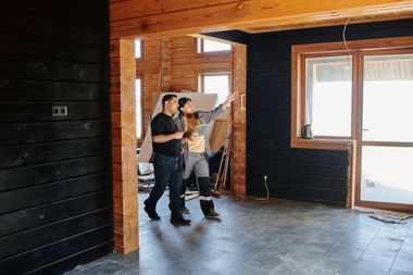 Two people inspecting a partially renovated room with exposed wood beams and bare concrete floors.