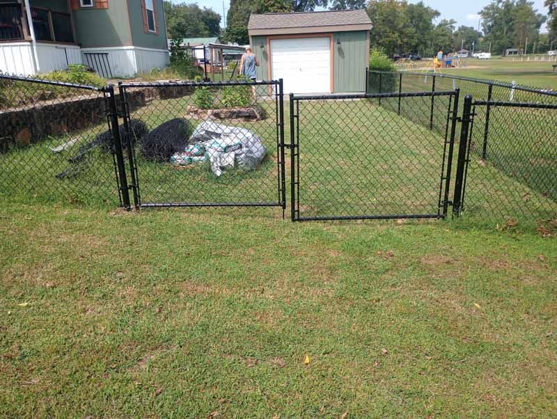 Black chain-link fence with gates encloses a grassy yard; a shed and house are in the background.