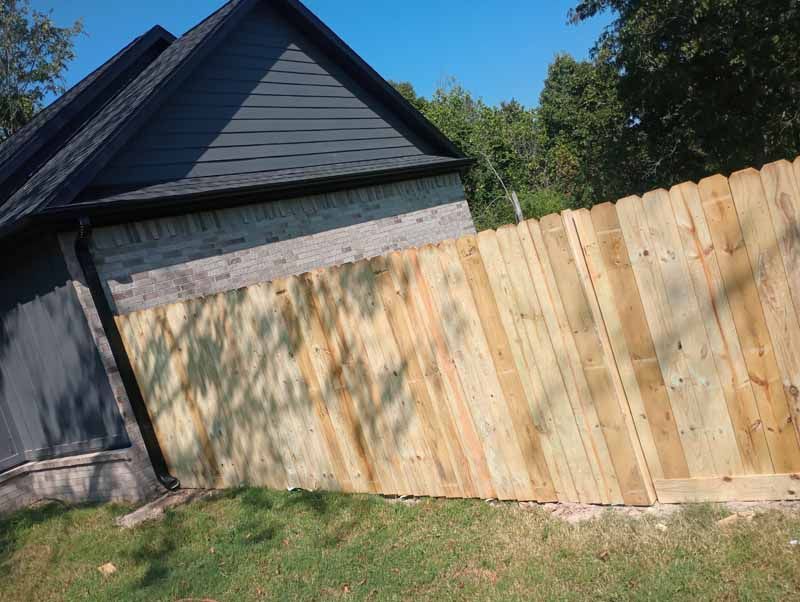 Wooden fence alongside a building with a dark roof and a grassy yard under a blue sky.