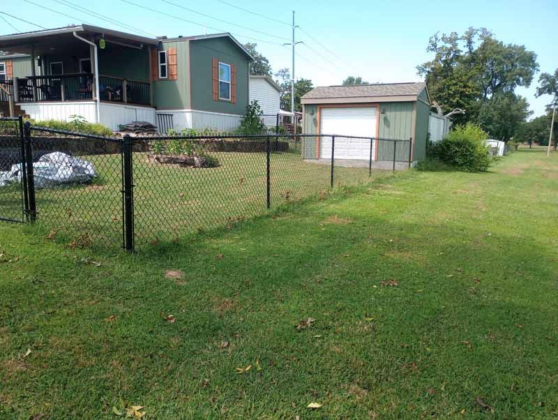 Black chain link fence encloses a green lawn, house, and small garage on a sunny day.