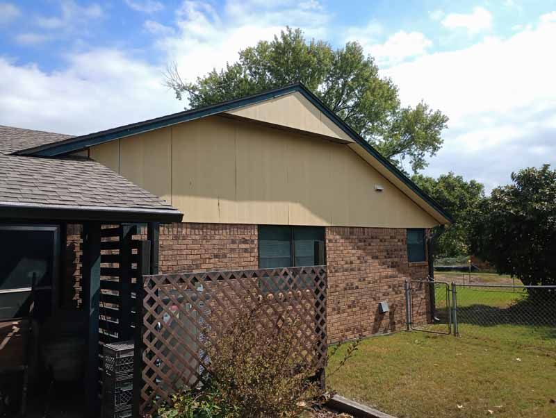 Side view of a brick house with tan siding, a dark roof, and a wooden fence in the yard.