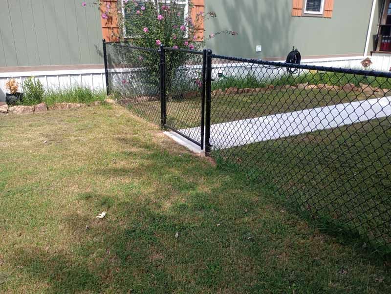 Black chain-link fence with gate in front yard with patchy grass, green house in background.
