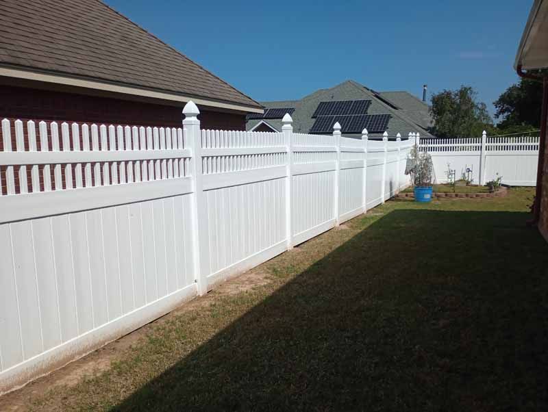 White vinyl fence around a backyard with green grass and brown roofed houses in the background.