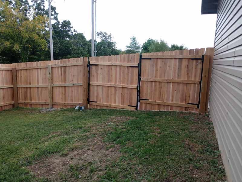 Wooden fence in backyard with gate, adjacent to a house. Green grass and trees visible.