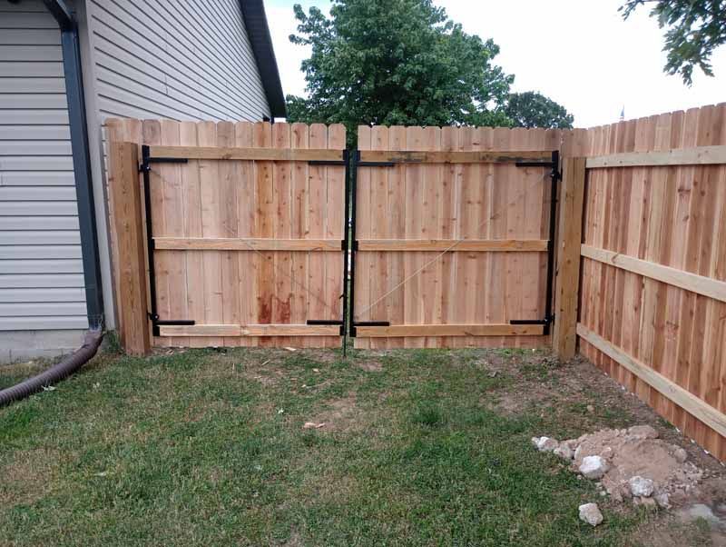 Wooden double gate in a wooden fence; surrounded by grass and a building.