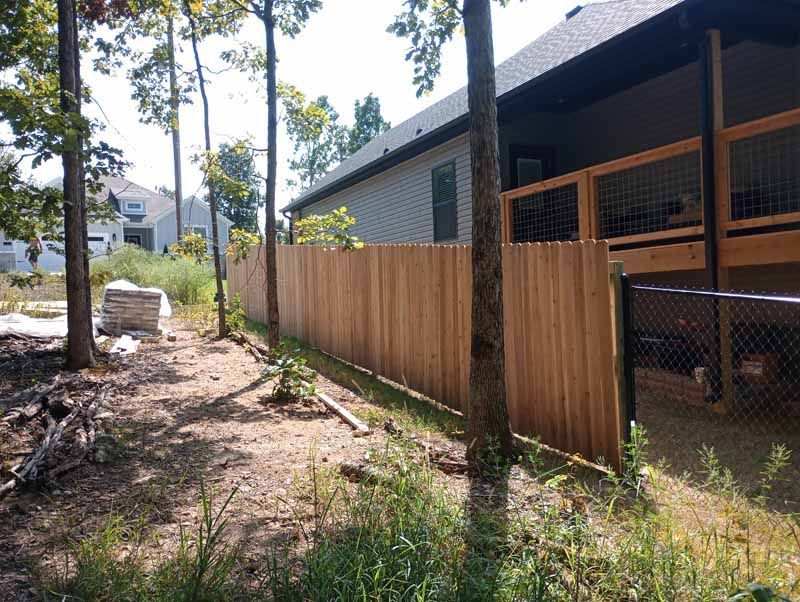 Wooden privacy fence alongside a house with a deck, trees in the yard.