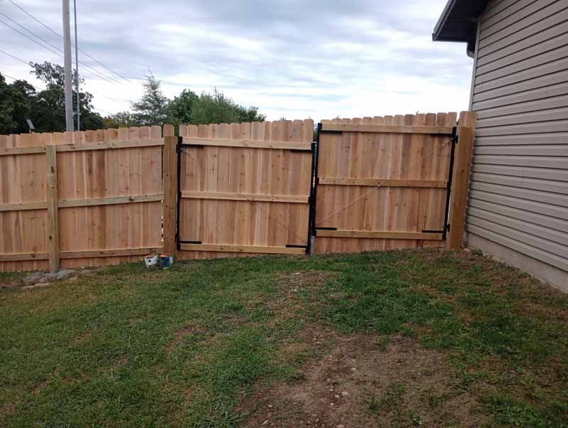 Wooden fence with black metal gate in grassy backyard, next to a beige house.