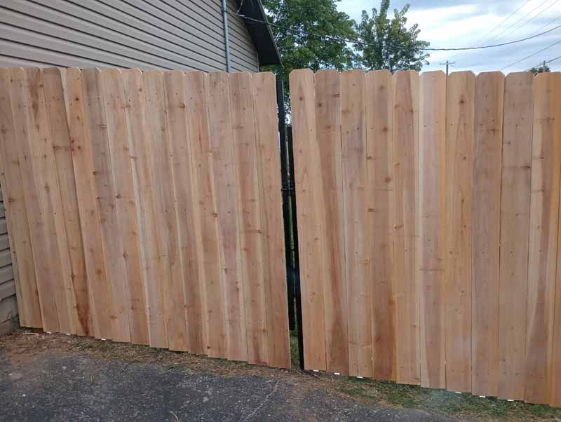 Wooden fence gate, closed, on a paved surface. Cedar planks are vertically aligned and frame a black metal hinge.