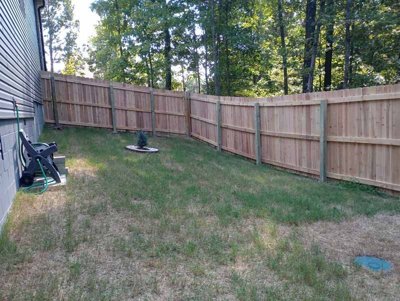 A wooden fence encloses a grassy backyard next to a house. Trees in the background.