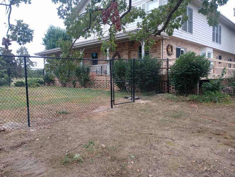 Black chain-link fence surrounds a house with a porch and wooden deck. Dry grass and shrubs are in the yard.