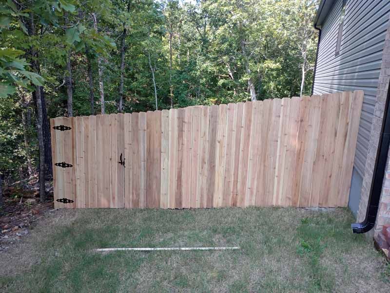 Wooden fence with gate next to a house and trees in the background, set on grass.