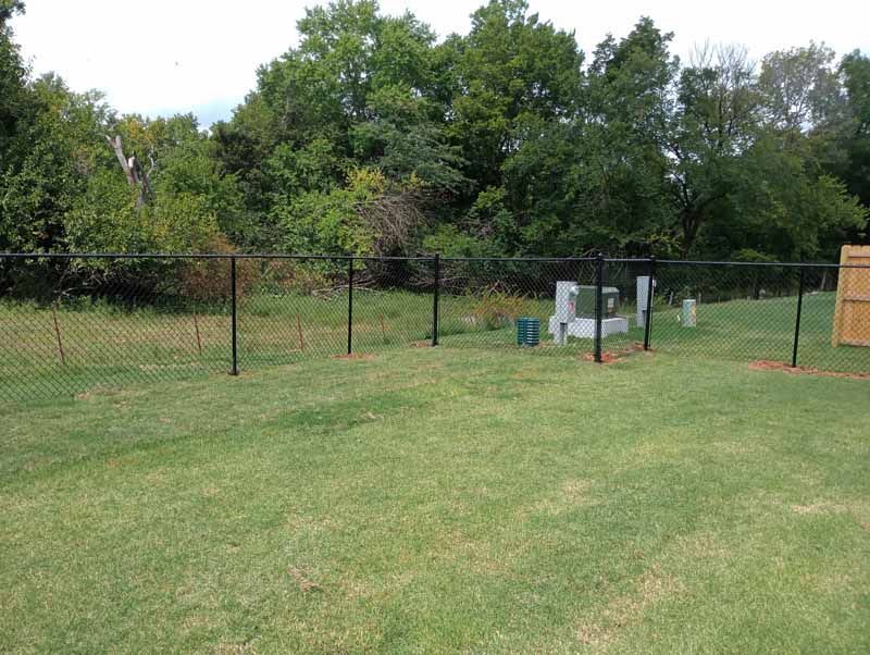 Black chain-link fence in backyard with trees in the background. Green grass covers the yard.