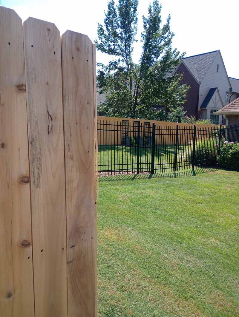 Wooden fence in foreground, green lawn, black wrought-iron fence, and houses in the background.