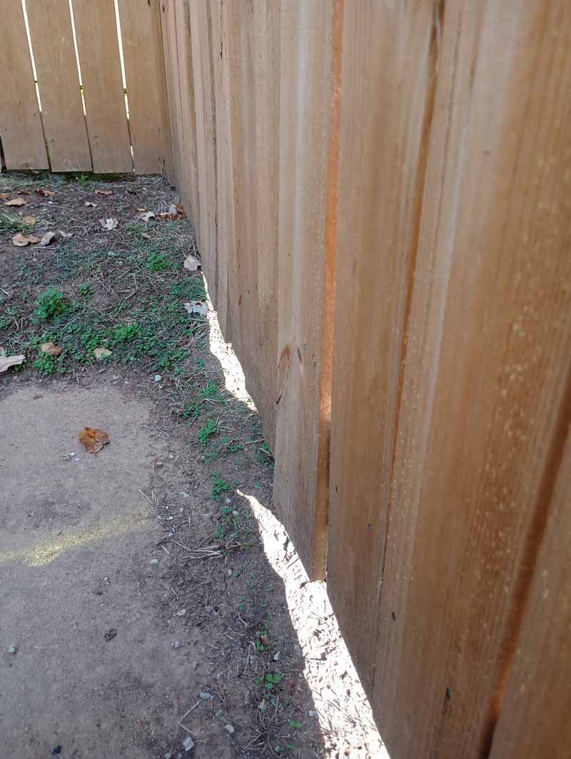 Wooden fence alongside a patch of grass and dirt. Sunlight casts a shadow on the ground.