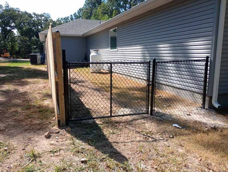 Black chain link fence with a gate, next to a wood fence and house with gray siding.