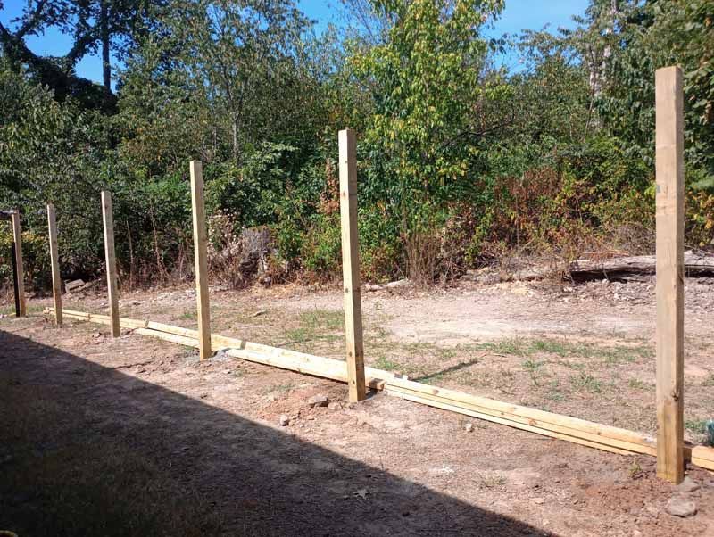 Wooden fence posts installed in a row, with horizontal boards laid out, set against a backdrop of trees.