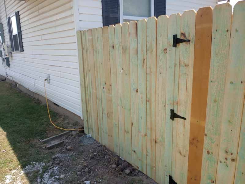 Wooden fence gate attached to a house; unfinished wood; black hinges.