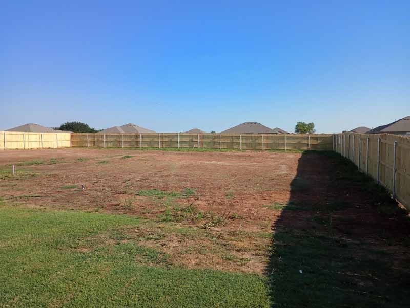 A mostly empty backyard with a wooden fence under a clear, blue sky.