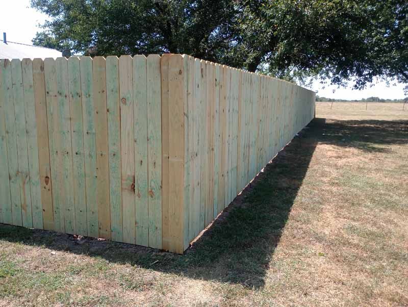 Wooden privacy fence in a grassy yard under a blue sky, casting a long shadow.