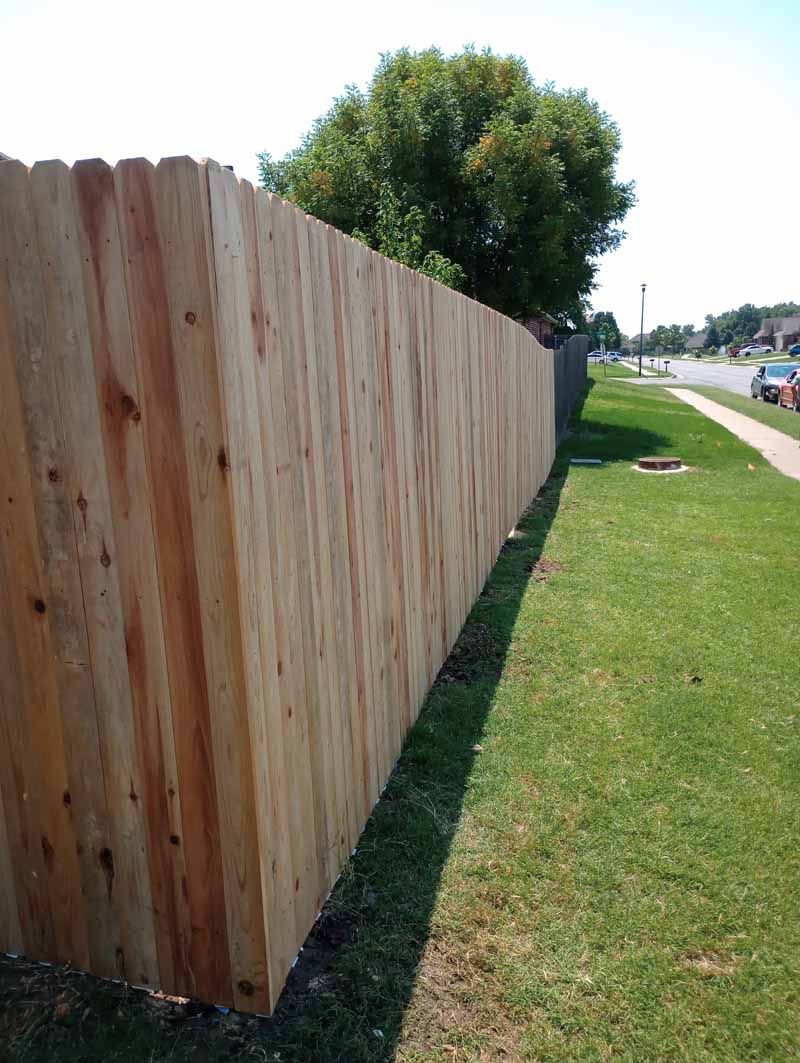 Wooden fence along a grassy lawn, with a tree and street visible in the background.
