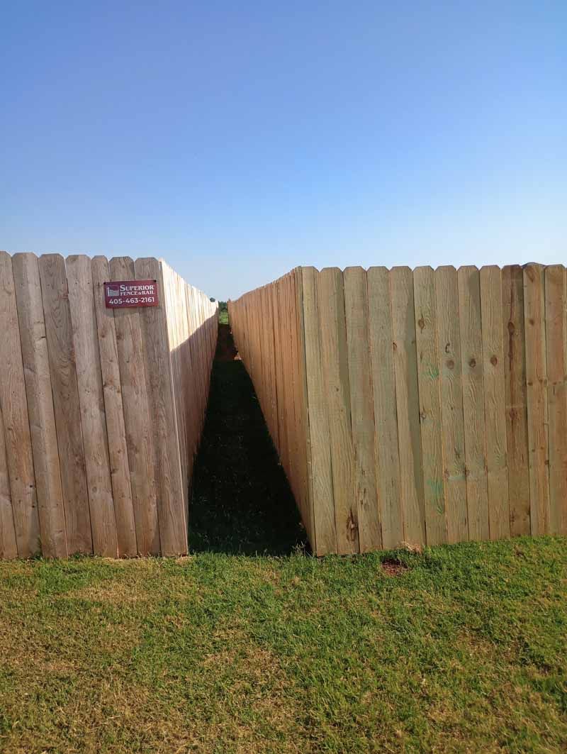 Wooden fence walls form a narrow, dark passageway through grass under a clear blue sky.