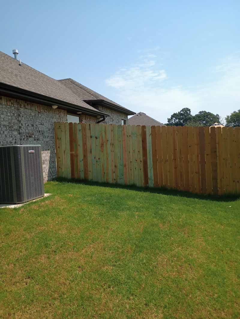 Backyard with wooden fence, grass, and air conditioning unit against a stone house under a blue sky.