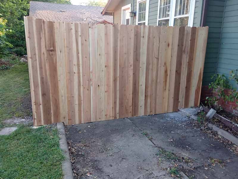 Wooden fence partially blocking the view of a house. It stands on a concrete slab and is bordered by grass.
