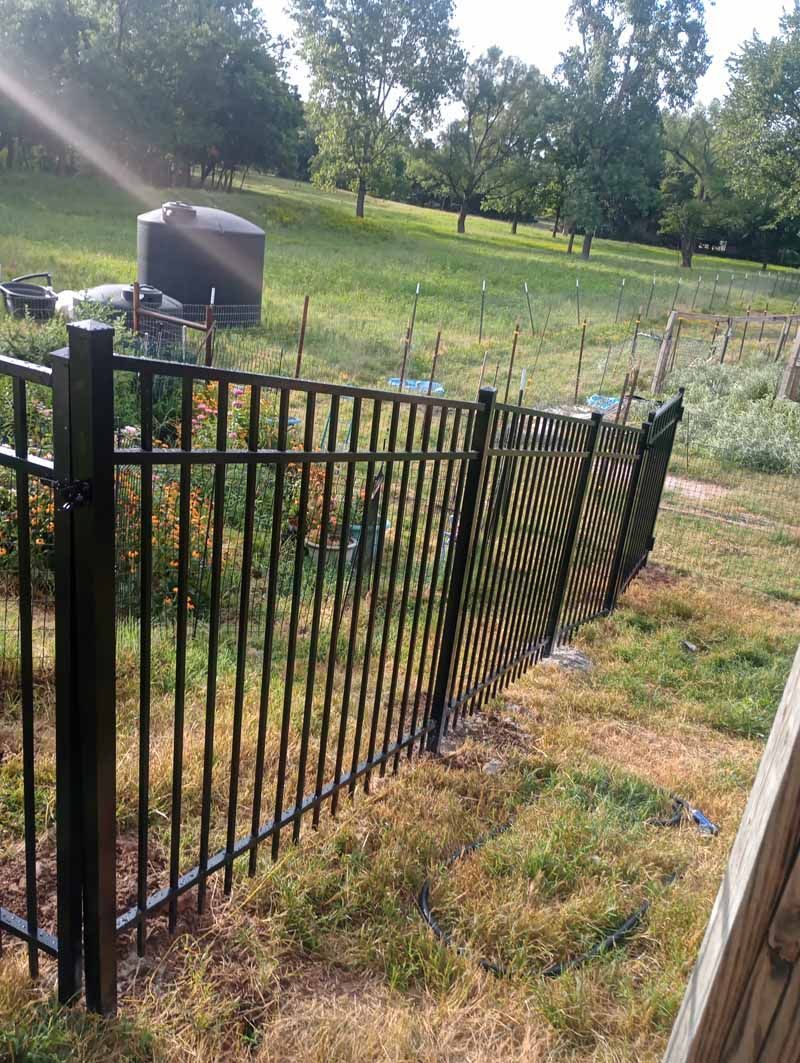 Black metal fence in a grassy yard, slightly curved, with a hillside and trees in the background.