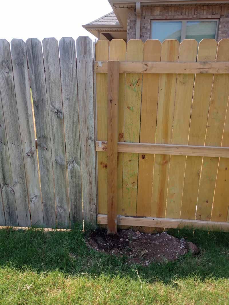 Two fences meet at a post: weathered gray wood and new, light brown wood. Earth mound at the base.