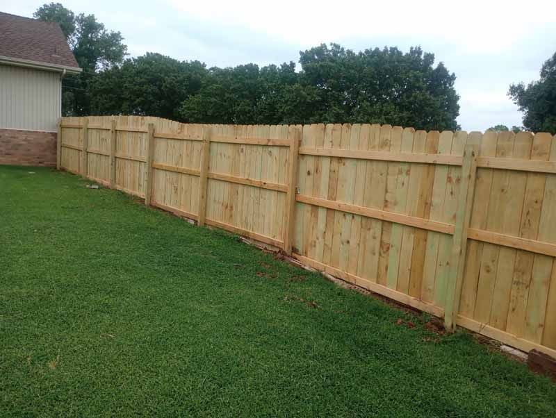 Wooden privacy fence along a grassy yard, with a house on the left and trees in the background.
