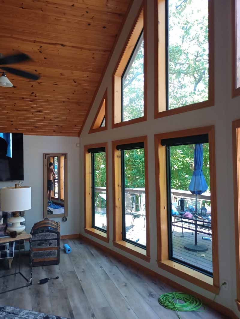 Interior room with tall windows overlooking a deck and trees, wood ceiling, light-colored walls and floor.
