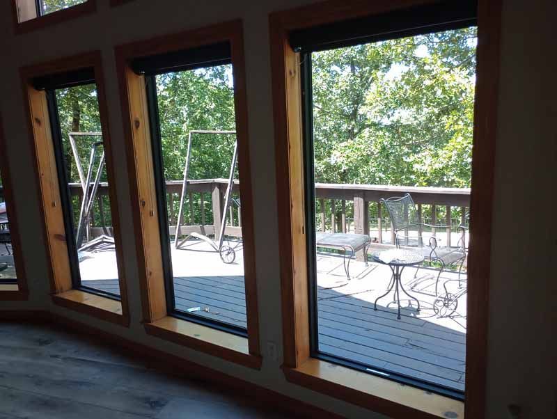 Three large windows with wooden frames overlook a deck and trees.