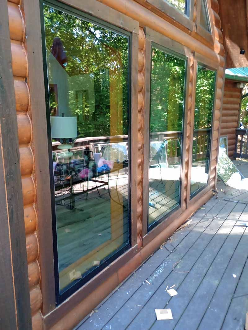 Log cabin exterior with three windows reflecting trees and a deck. Brown logs, green trees, blue sky.