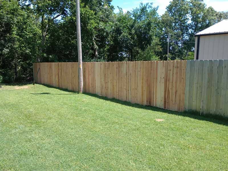 Wooden fence bordering a grassy yard with trees and a building in the background on a sunny day.