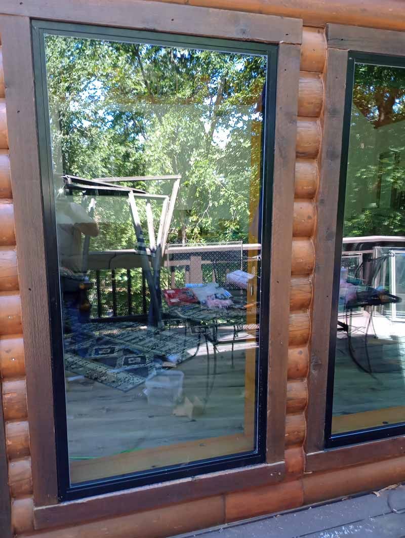 Exterior view of a window reflecting a wooden deck, trees, and sky. The window is framed in brown, set in a log cabin wall.