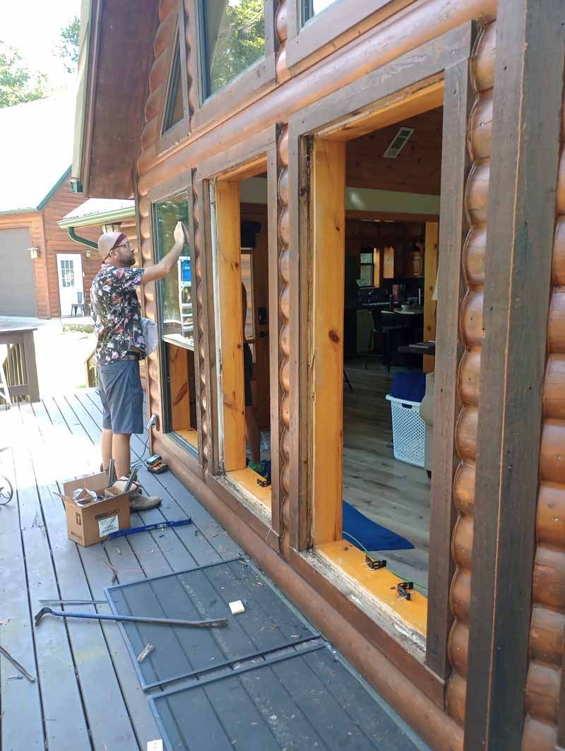 Man replacing window in a log cabin. He's on a wooden deck. The cabin is brown with large logs.