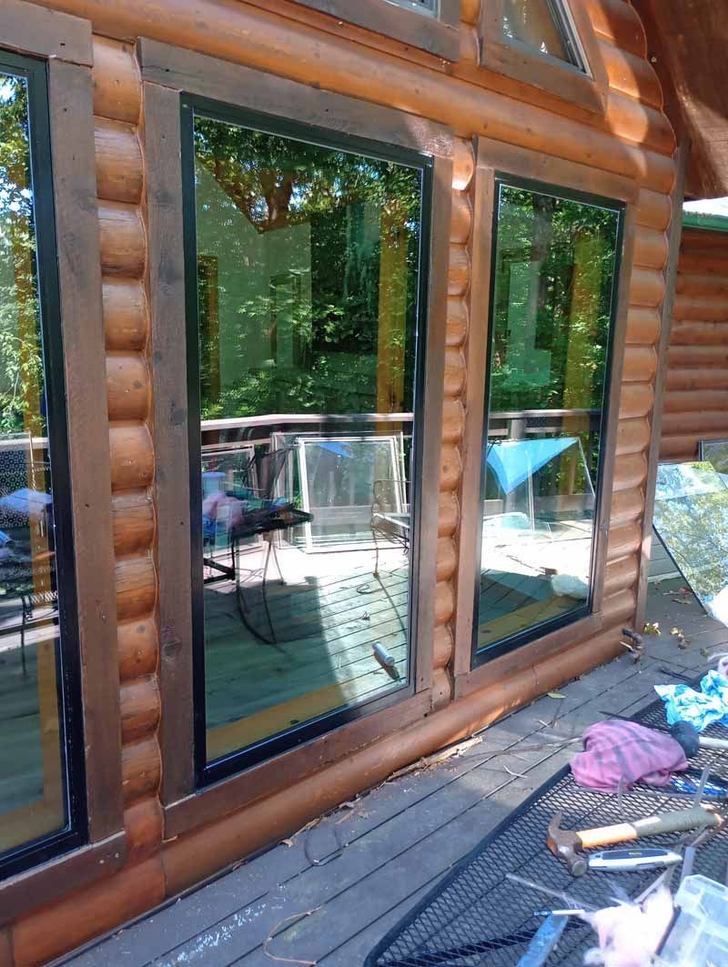 Three tall windows with brown frames in a log cabin wall, reflecting outdoor trees and a deck.