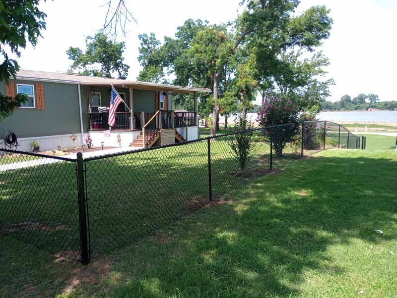 Green house with porch, black chain link fence, and American flag on a grassy lawn near water.