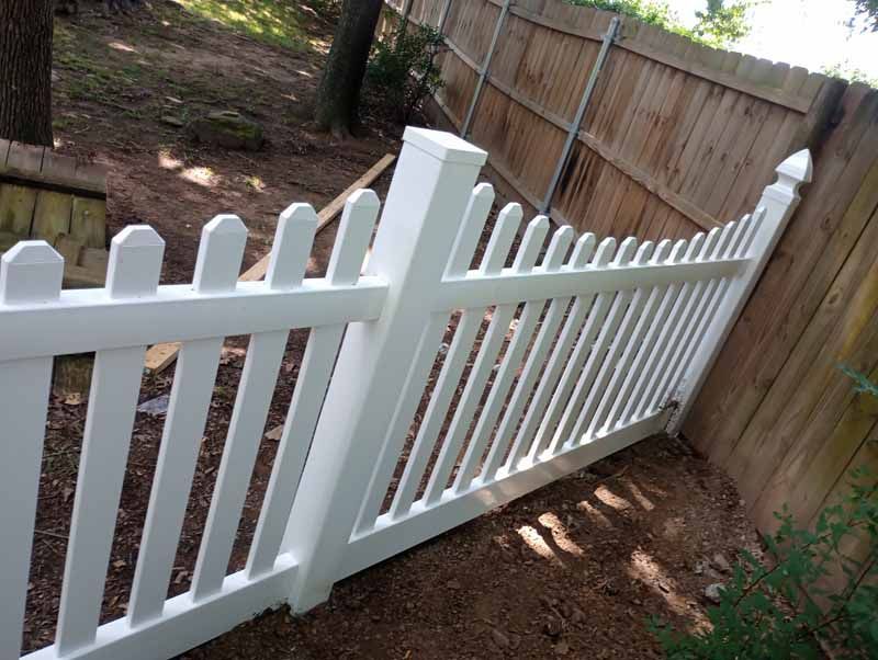 White picket fence next to a wooden privacy fence in a yard with trees.