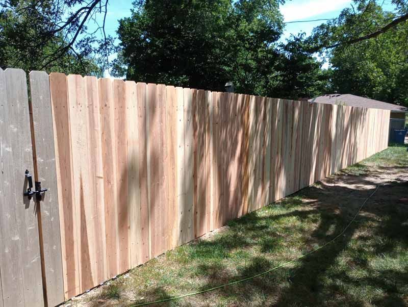 Wooden privacy fence in a yard with a gate, trees and a building in the background.