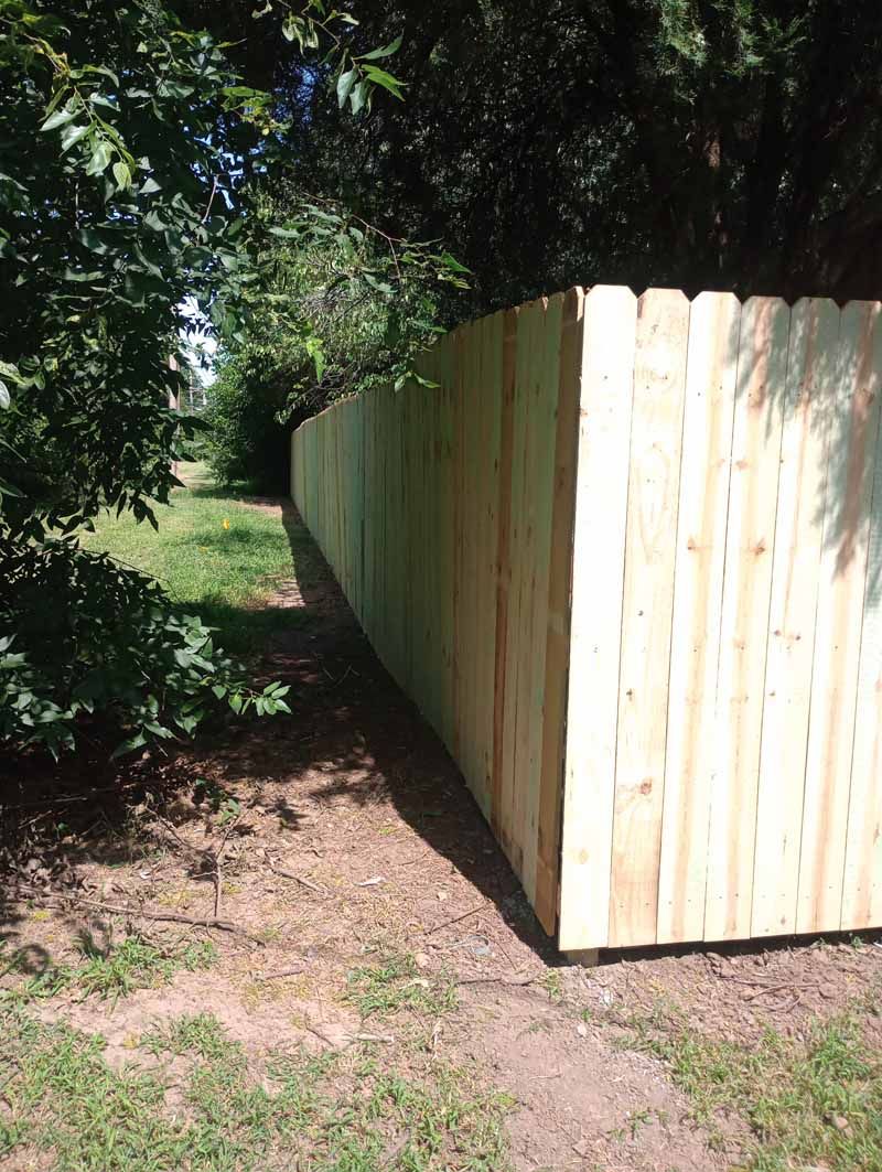 Wooden fence along a grassy path, surrounded by trees and sunlight.