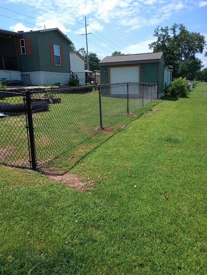 A chain-link fence encloses a grassy area next to a green building and a shed.