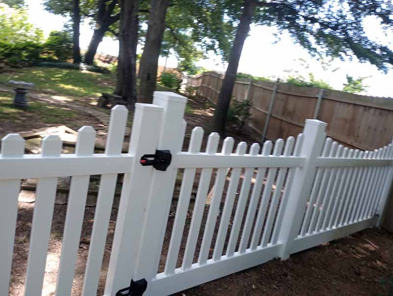 White picket fence gate in a backyard with trees and a wooden fence in the background.