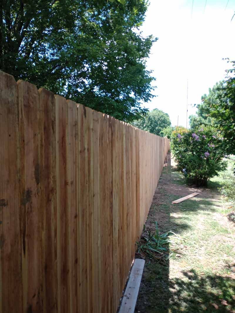 Wooden fence in a yard, extending towards the distance. Sunlight, trees, and grass are visible.